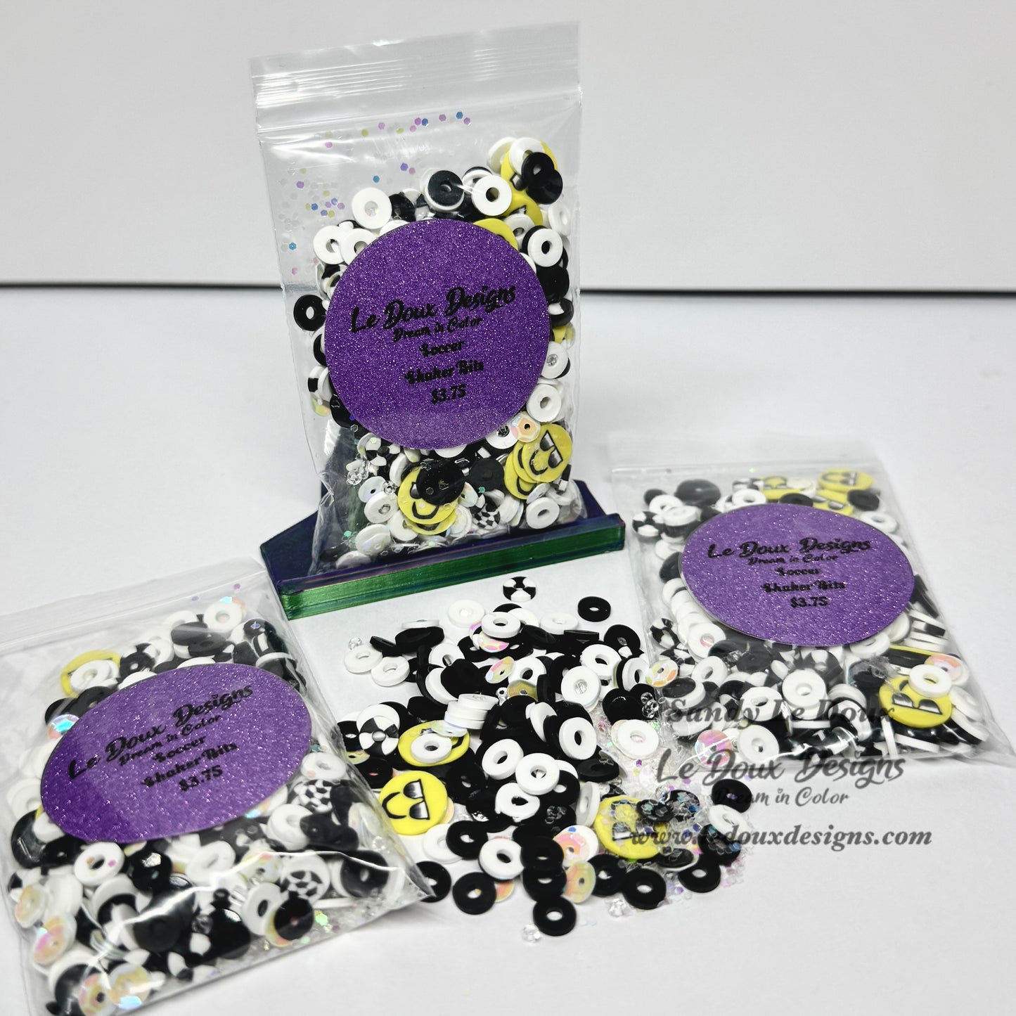 Three clear plastic bags of decorative craft beads with purple glitter round labels, plus a small pile of loose beads in the center, photographed on a white background. The beads are primarily black and white with some yellow smiley-face style accents.
