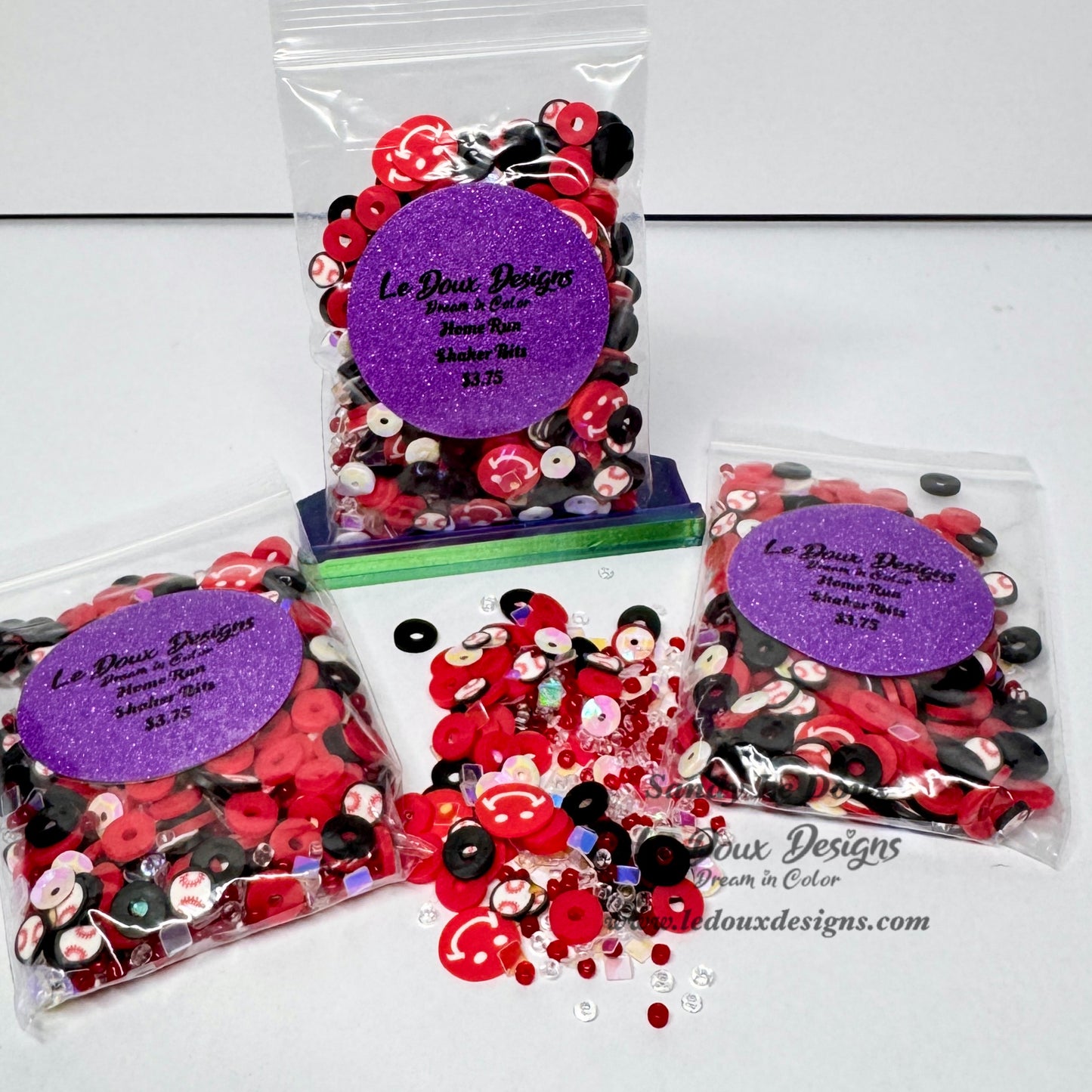Three clear plastic bags filled with red, black, and white decorative buttons and sequins, arranged on a white background. Each bag has a purple glitter label reading 'Le Doux Designs'.