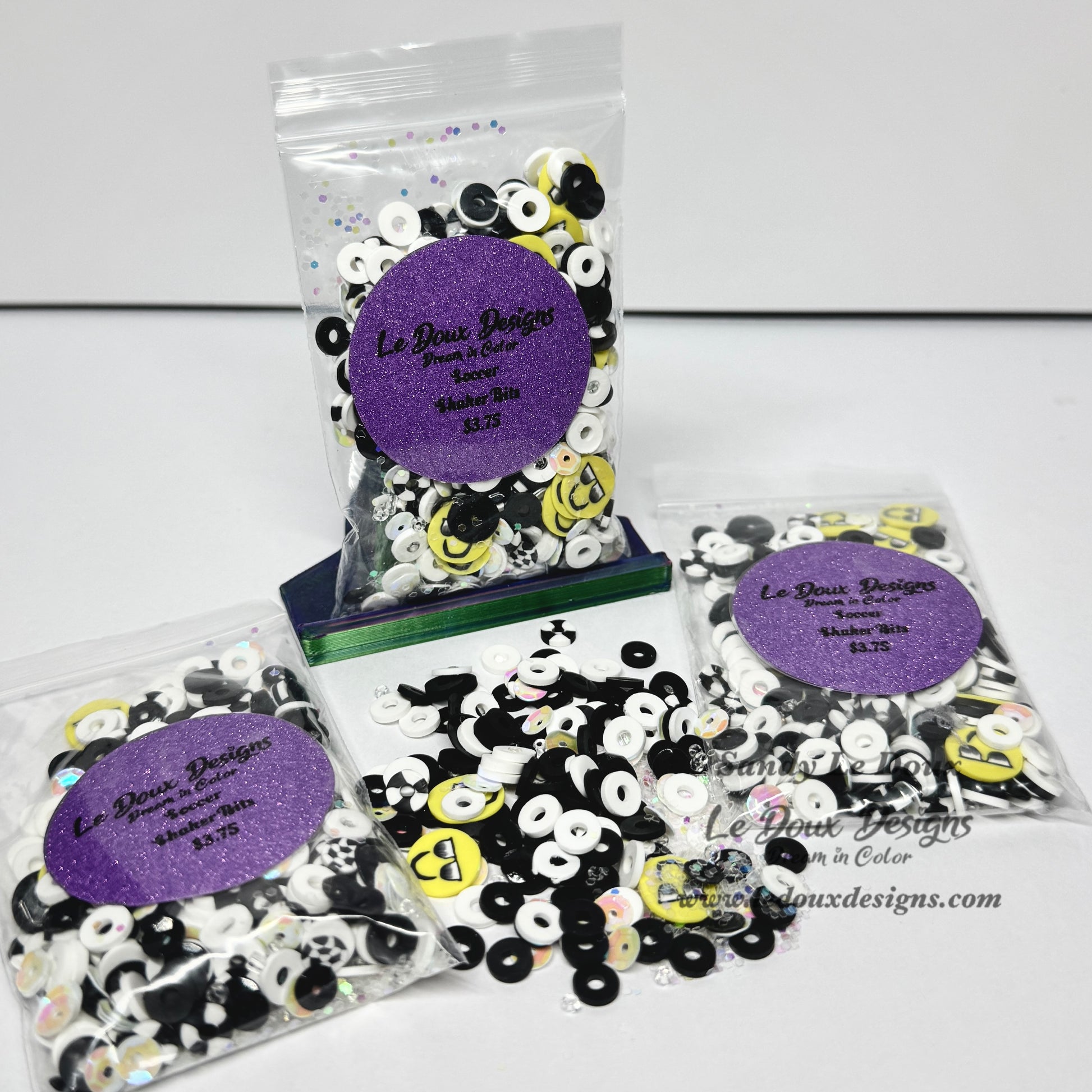 Three clear plastic bags of decorative craft beads with purple glitter round labels, plus a small pile of loose beads in the center, photographed on a white background. The beads are primarily black and white with some yellow smiley-face style accents.